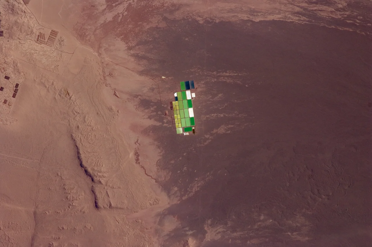 Brightly coloured solar evaporation (salt) ponds in a desert landscape give this astronaut photo an unreal quality. The ponds sit near the foot of a long alluvial fan in the Pampa del Tamarugal, the great hyper-arid inner valley of Chile’s Atacama Desert. The alluvial fan sediments are dark brown, and they contrast sharply with tan sediments of the Pampa del Tamarugal. Nitrates and many other minerals are mined in this region. A few extraction pits and ore dumps are visible at upper left. Iodine is one of the products from mining; it is first extracted by heap leaching. Waste liquids from the iodine plants are dried in the tan and brightly coloured evaporation ponds to crystallize nitrate salts for collection. Coordinates: -20.946162, -69.514961
This image was aquired with a Nikon D2X digital camera fitted with a 400 mm lens, and is provided by the ISS Crew Earth Observations experiment and Image Science &amp; Analysis Laboratory, Johnson Space Centre.
