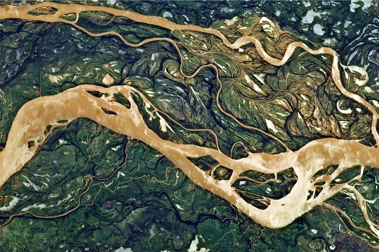 This astronaut photograph shows a 29-kilometre stretch of the Paraná, downstream of the small city of Goya, Argentina (just off the top left of the image).
The Paraná River ranges up to 3 kilometres wide along the reach illustrated in this image. The main channel is deep enough to allow smaller ocean-going ships to pass north to the capital city of Asunción, Paraguay, fully 1,200 kilometres inland and well out of the image.
The river’s dark brown shading indicates a heavy load of muddy sediment; smaller side channels also carry this mud. Numerous lakes are typical on active floodplains, and appear here as irregular bodies of water. Some appear brown, indicating that they probably have been refilled during recent rises and floods of the active channels.
The Paraná floodplain occupies the entire image; it is so wide—18 kilometres in this view—that its banks are not visible. Numerous curved, meandering channels are the most prominent characteristic of the floodplain, indicating prior positions of the river and its channels. As riverbeds move laterally by natural processes, they leave remnants of their channels, which appear as lakes and finally fill with mud. This is an excellent image for illustrating these meander forms.
From a geological standpoint, it is interesting that almost all of the old channels are similar in curvature to today’s side channels. However, almost none of them seem to show prior positions of the main, wide Paraná channel.