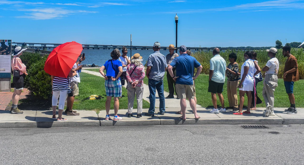In the foreground, an asphalt road can be seen. An NPS Ranger can be seen presenting a piece of paper while giving an interpretive talk to a group of 13 guests standing on a sidewalk at historic Long Wharf in Cambridge, MD. A light post, the Choptank River, and the Choptank Bridge can be seen in the background.
An NPS ranger gives an interpretive talk at historic Long Wharf in Cambridge, MD.
Keywords: Harriet Tubman; Harriet Tubman Underground Railroad National Historical Park; ranger program; walk; photography