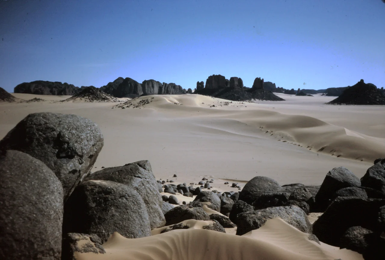 Location: On the southeastern edge of Tassili n'Ajjer, northwest of Djanet
The prevailing wind in the Sahara is the northeast trade wind (Harmattan). During the early Quaternary period, there were times when it blew much stronger than it does today. The sand in this picture was probably blown at that time from the Erg Ubari and Erg Murzouk areas in southwestern Libya across the gently ascending plateau of the Tassili n'Ajjer and deposited here, in the lee behind the mountain ridge.
The rounded boulders in the foreground are the result of insolation weathering. The large temperature differences between day and night build up tension in the rock, especially on protruding parts. These splinter off, and as the boulder becomes smaller and smaller, its shape becomes more and more rounded.