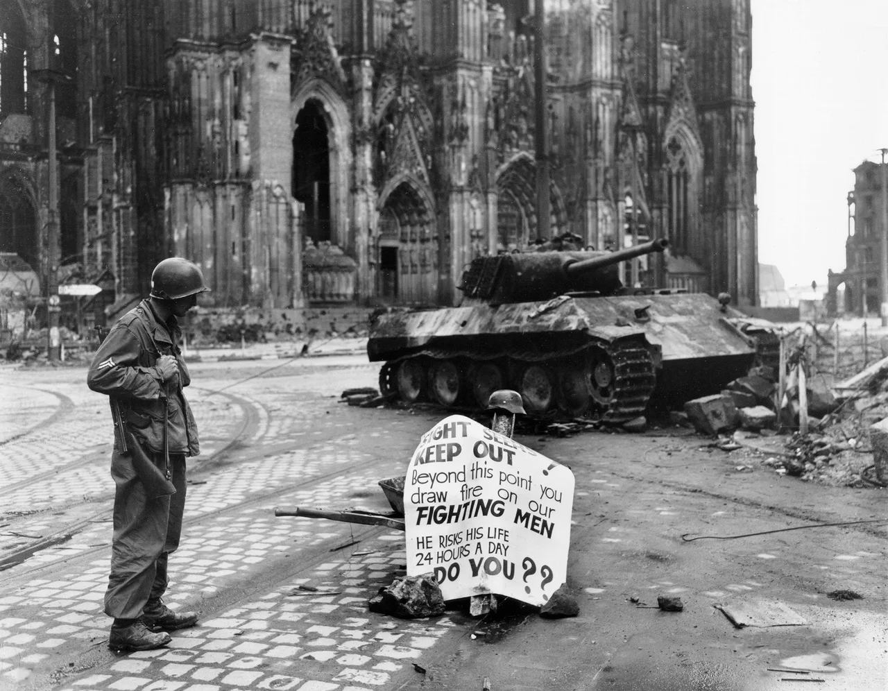 Original caption: Cologne, Germany - Cpl. Luther E. Boger, Concord, N.C., skytrooper, reads a warning sign in the street. This street leads to the Rhine River and is under observation of the Germans who occupy a stronghold there. Cpl. Boger is with the 82nd Airborne Division. 4 April 1945. The German tank is burnt out and the torsion-bars have been destroyed by the immense heat of the fire.

Allied troops captured the western part of Cologne on 6th-7th March 1945. The German army still held the eastern shore of the Rhine and attacked the Allies with artillery. The rest of Cologne was captured between 12th and 15th April 1945. Until the 16th April a strip of about 500m along the shore had been declared as a restricted area and the cathedral was just within this zone.

Text on sign reads:

SIGHT SEERS
KEEP OUT!
Beyond this point you
draw fire on our
FIGHTING MEN
HE RISKS HIS LIFE
24 HOURS A DAY
DO YOU??